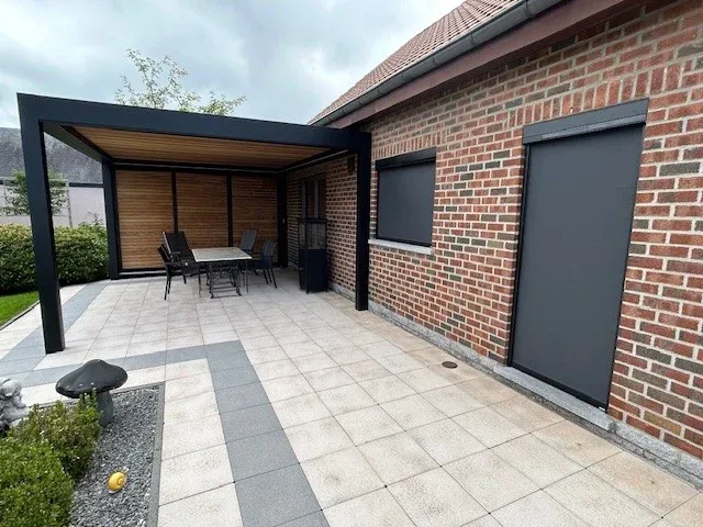 Terrasse en dalles claires devant une pergola anthracite à plafond bois, maison en briques rouges
