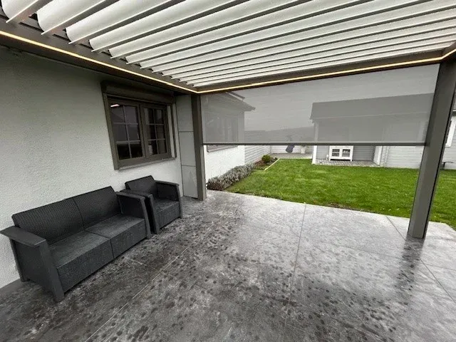 Pergola bioclimatique claire adossée à une maison, screens latéraux descendus et salon de jardin sur terrasse en béton