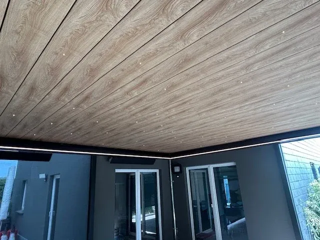 Plafond aspect bois d’une pergola, spots encastrés et bandeau LED, au-dessus d’une façade anthracite